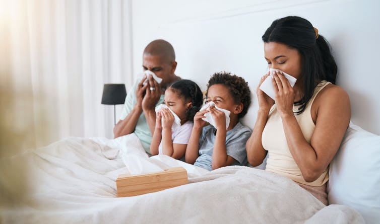 A family, mom, dad and two young children all sit in bed together blowing their noses.