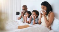 A family, mom, dad and two young children all sit in bed together blowing their noses.