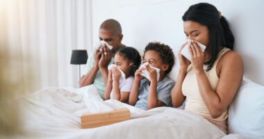 A family, mom, dad and two young children all sit in bed together blowing their noses.