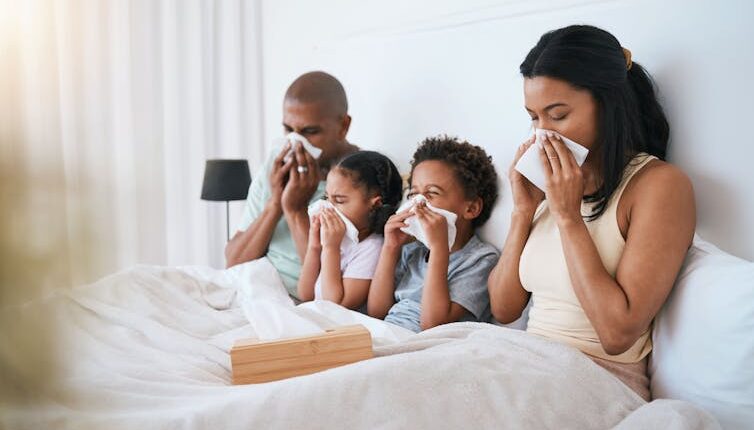A family, mom, dad and two young children all sit in bed together blowing their noses.