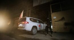 A gunman wearing the uniform of the al-Qassam Brigades, the military wing of Hamas, stands guard as Red Cross vehicles enter a warehouse allegedly to collect coffins containing the bodies of four deceased hostages, in Gaza.