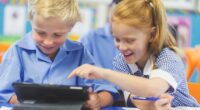 Students using a digital tablet in a classroom. There is a boy and a girl, both are wearing school uniforms and are happy and smiling.