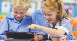 Students using a digital tablet in a classroom. There is a boy and a girl, both are wearing school uniforms and are happy and smiling.