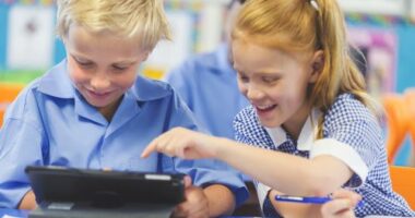 Students using a digital tablet in a classroom. There is a boy and a girl, both are wearing school uniforms and are happy and smiling.