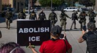 Protesters gather in front of ICE officers. One holds a sign reading ABOLISH ICE