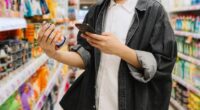 Asian Chinese Student Looking At Smartphone While Choosing Items In Supermarket.