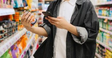 Asian Chinese Student Looking At Smartphone While Choosing Items In Supermarket.