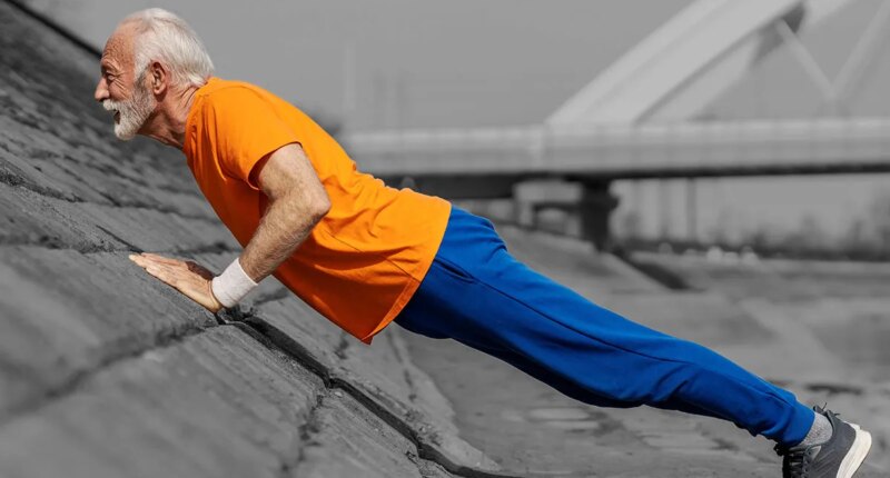 Profile view of an old athlete doing pushups and strength exercises on river shore.