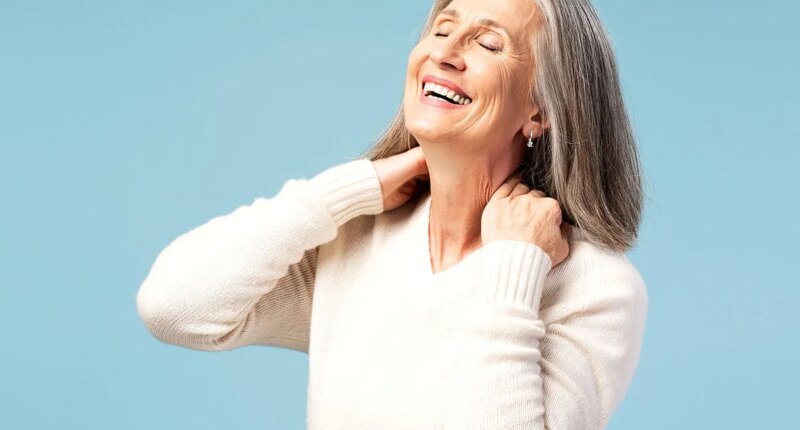 Portrait of smiling senior gray haired woman touching neck, having pain with closed eyes fashion model posing isolated on blue background. Health care concept