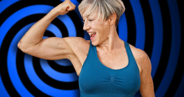 A fit, mature woman with short grey hair flexes her muscles against a pink isolated background, displaying strength and vitality.