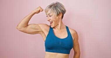 A fit, mature woman with short grey hair flexes her muscles against a pink isolated background, displaying strength and vitality.