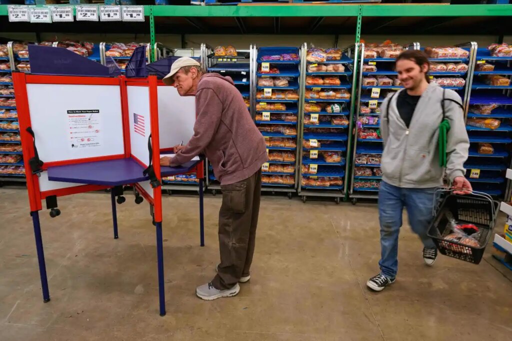 Bob Walser votes while a shopper walks past at the Checkers grocery store in Lawrence, Kan., Tuesday, Nov. 4, 2025. (AP Photo/Charlie Riedel)