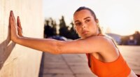 Focused woman in orange sports top leaning against a wall exercising outdoors