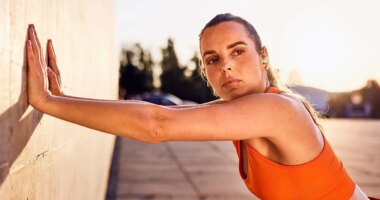 Focused woman in orange sports top leaning against a wall exercising outdoors