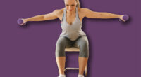 Smiling muscular woman in shape sitting on the chair at home and doing fitness exercises for arms with dumbbells.