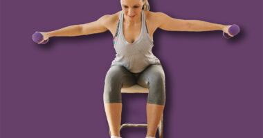 Smiling muscular woman in shape sitting on the chair at home and doing fitness exercises for arms with dumbbells.