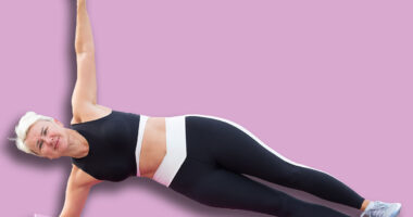 Athletic woman is engaging in a side plank exercise on a pink yoga mat, with sandy beach and palm trees creating a serene atmosphere