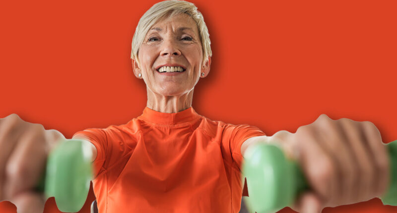 An elderly woman sits on a chair, lifting green dumbbells while smiling. She exercises in a bright living room with plants, promoting strength, fitness, and an active lifestyle.