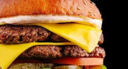 A mouthwatering burger features two juicy beef patties layered with cheddar cheese, fresh lettuce, sliced tomatoes, pickles, and creamy sauce, all in a toasted sesame seed bun.