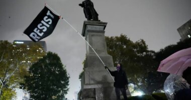 A protestor waves a flag in a pouring rain during a demonstration against an impending Customs and Border Patrol immigration crackdown in New Orleans, Monday, Dec. 1, 2025. (AP Photo/Gerald Herbert)