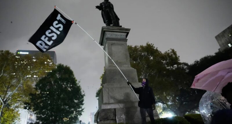 A protestor waves a flag in a pouring rain during a demonstration against an impending Customs and Border Patrol immigration crackdown in New Orleans, Monday, Dec. 1, 2025. (AP Photo/Gerald Herbert)