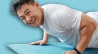 Active asian mature man standing in plank pose or doing push ups on mat at home and smiling at camera. Active korean male leading active lifestyle, exercising indoors