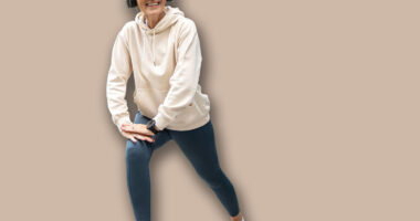 Smiling senior woman in headphones stretching her legs on a paved walkway, exercising outdoors in an urban environment with modern buildings in the background, promoting active aging and well-being