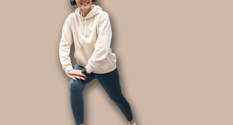 Smiling senior woman in headphones stretching her legs on a paved walkway, exercising outdoors in an urban environment with modern buildings in the background, promoting active aging and well-being
