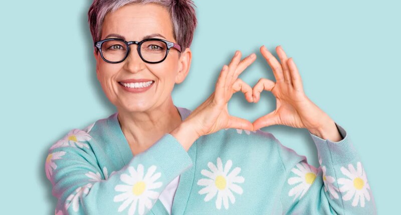 Charming senior woman smiling in colorful cardigan making heart gesture against pink background