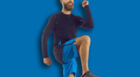 Male athlete in modern activewear having his routine fitness workout at home. Happy handsome bearded athletic man in sports shorts doing high knees walk in place exercise on rubber mat in living room