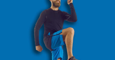 Male athlete in modern activewear having his routine fitness workout at home. Happy handsome bearded athletic man in sports shorts doing high knees walk in place exercise on rubber mat in living room