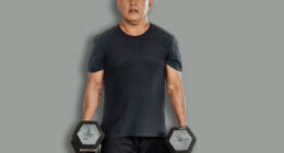 Middle-aged Southeast Asian man carries two dumbbells in a farmer’s walk for forearm or grip strength and endurance training inside open-air gym.