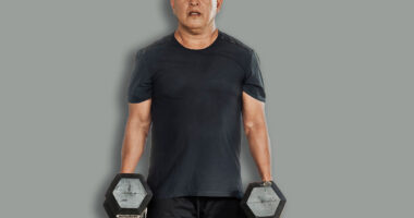 Middle-aged Southeast Asian man carries two dumbbells in a farmer’s walk for forearm or grip strength and endurance training inside open-air gym.