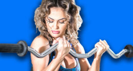 Attractive woman with curly hair doing preacher curl biceps exercise on bench with EZ curl bar in modern fitness center. Toned image.