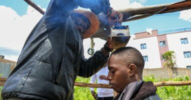 Barber and viral content creator Safari Martins, demonstrates one of his inventive shaving methods using an iron box while grooming Ian Njenga in Kiambu, Kenya, Wednesday, Nov. 26, 2025. (AP Photo/Andrew Kasuku)