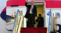 British Prime Minister Keir Starmer waves from the top of an airplane's red-carpeted stairs.