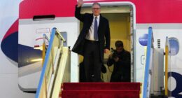 British Prime Minister Keir Starmer waves from the top of an airplane's red-carpeted stairs.