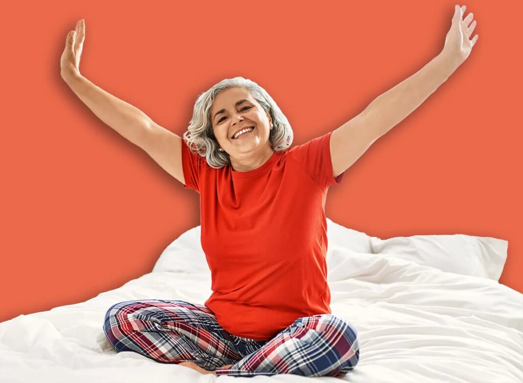Middle age grey-haired woman smiling happy sitting on the bed with hands raised up at bedroom.