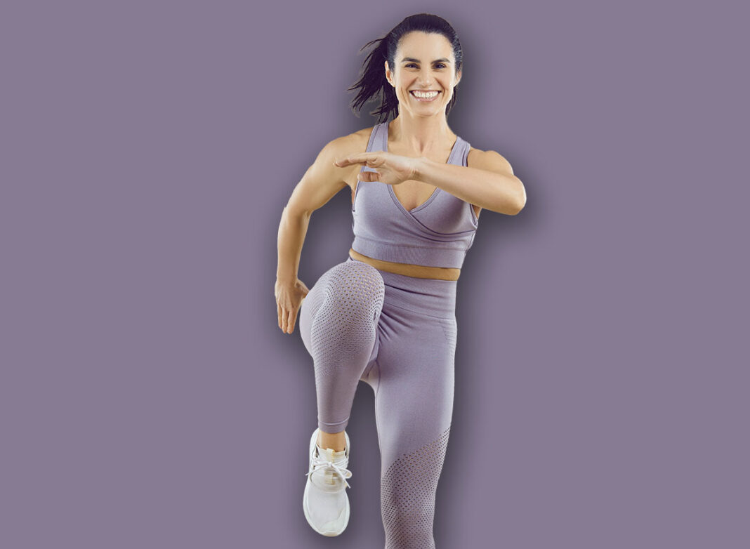 Cheerful sportswoman performs sport jumps raising her knees high on beige background. Full length portrait of smiling energetic woman in sportswear showing sports exercises. Banner. Isolated.