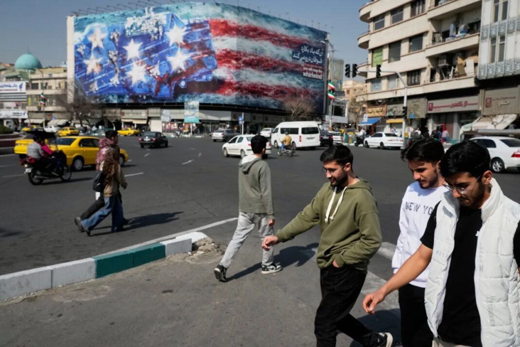 Pedestrians walk past a billboard depicting a U.S. aircraft carrier with damaged fighter jets on its deck and a sign in Farsi and English reading, "If you sow the wind, you