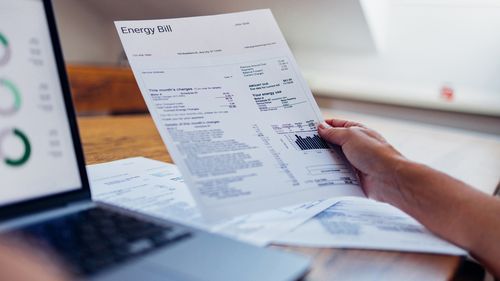 A person reviews their energy bill while using a laptop at home. The document includes various charges and electricity usage information.