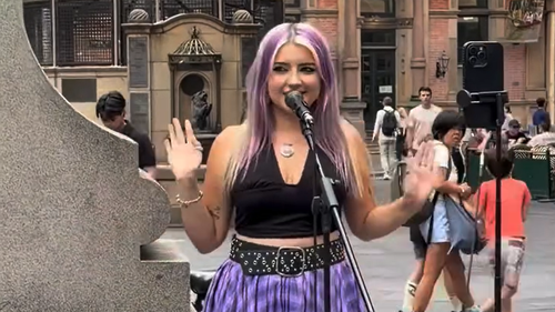 A young woman with purple hair sings into a microphone while busking on Sydney's George Street. She's wearing a black top and purple skirt.