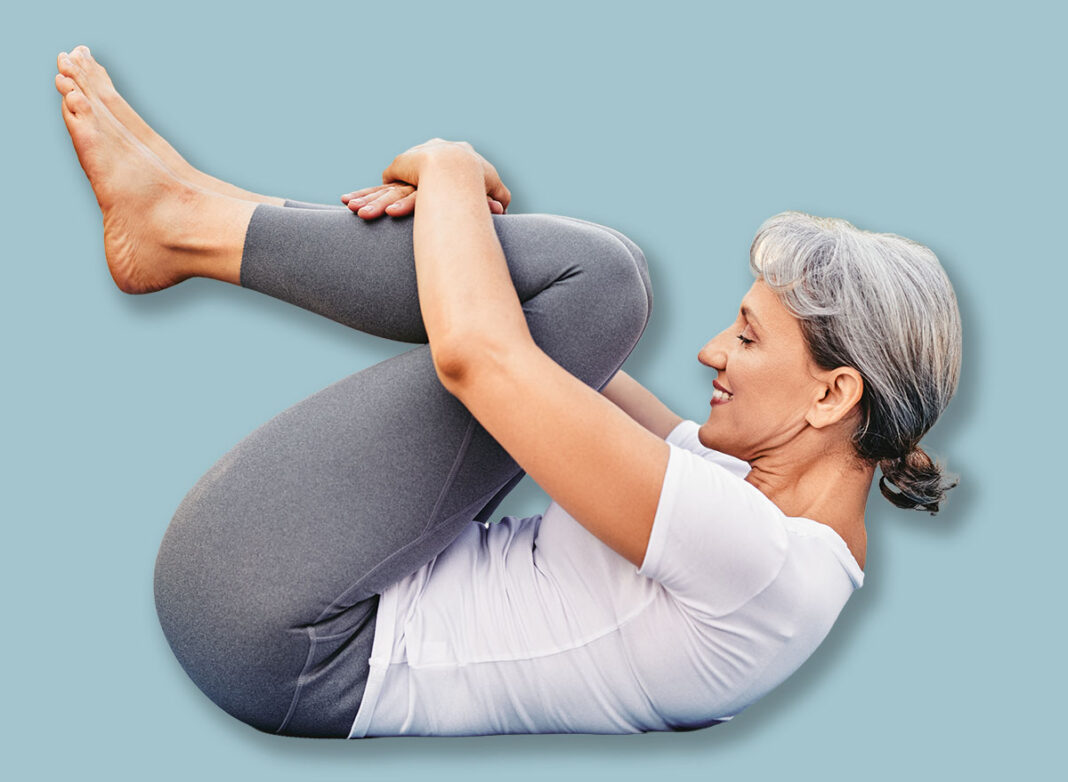 Side view of mature woman in sports wear lying on mat hugging her knees to chest after yoga practice outdoor in park, healthy active senior female doing stretching exercises in morning on fresh air