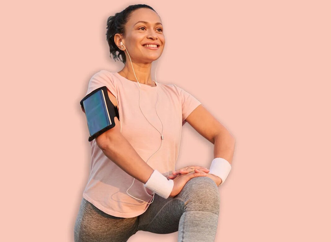 Beautiful middle aged sportswoman with earphones and smartphone holder enjoying morning workout at sunrise, stretching legs, doing lunges, squatting before jogging. Female athlete working out outdoor