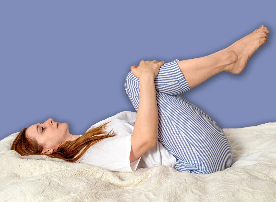 Woman doing exercises in bed at home in the morning. Apanasana Yoga pose knees-to-chest body stretch. Lady with long hair in pajamas. Girl stretches her muscles on a clean white bedding