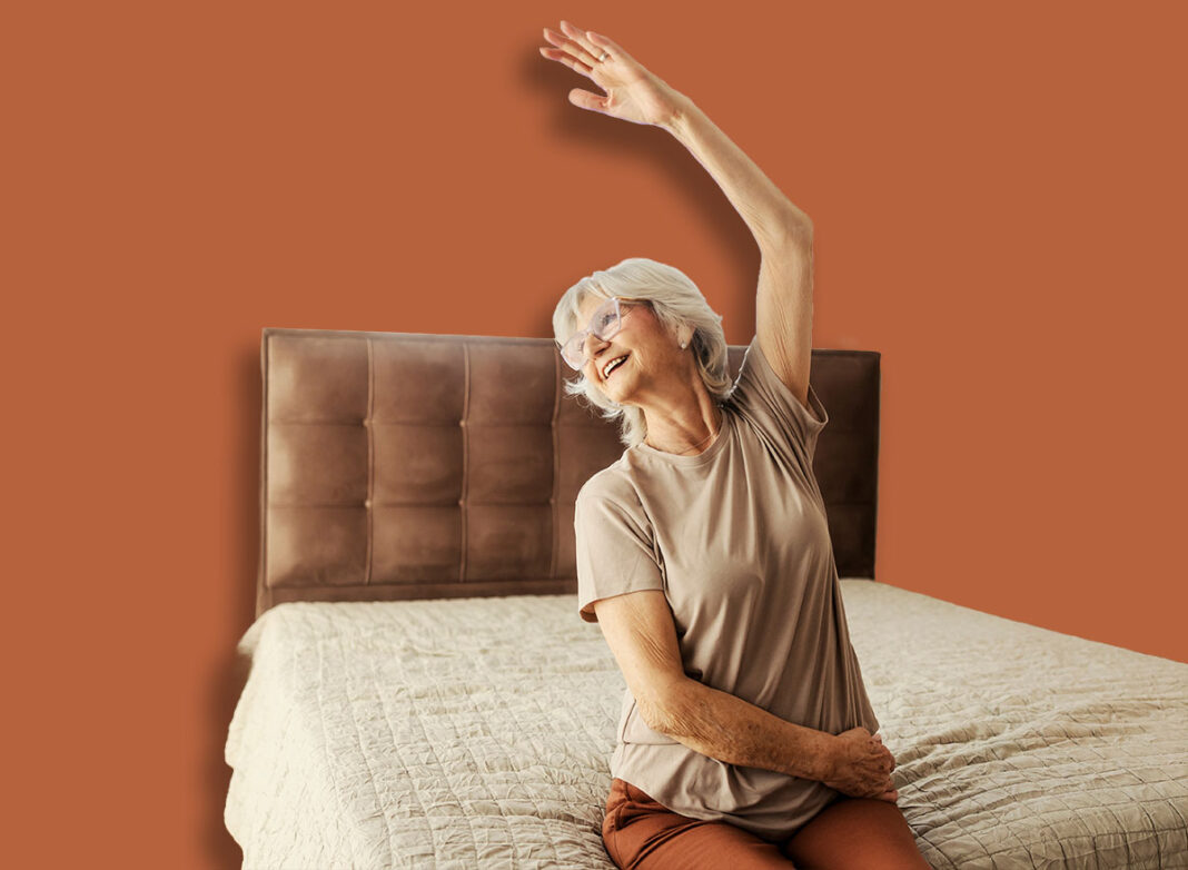 A happy senior woman sitting on a bed at home and doing senior fitness exercises.