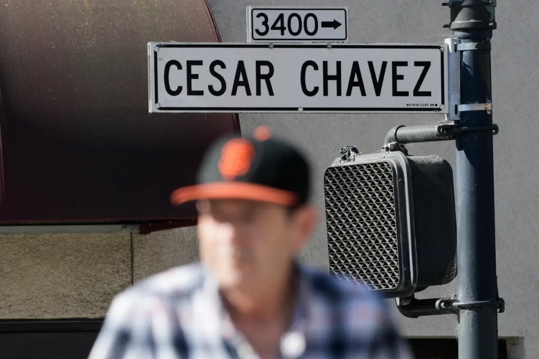 A pedestrian walks below a César Chavez Street sign in San Francisco, Wednesday, March 18, 2026. (AP Photo/Jeff Chiu)