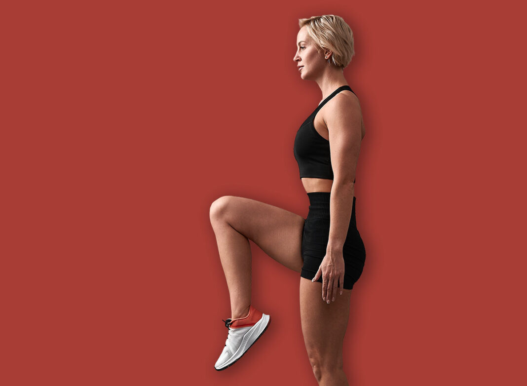 Standing exercise lifting leg with bent knee hands lowered pressed against the body. A young woman of athletic build is engaged in fitness.