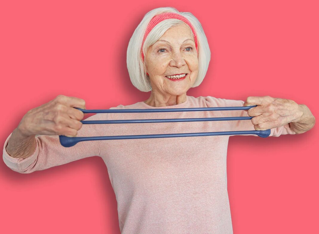 Happy elderly female exercising with rubber expander to strengthen her arm muscles on blurred background of flat