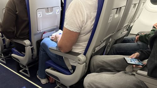 Passengers seated inside an airplane cabin with visible seat belts, legroom, and personal belongings during a commercial flight